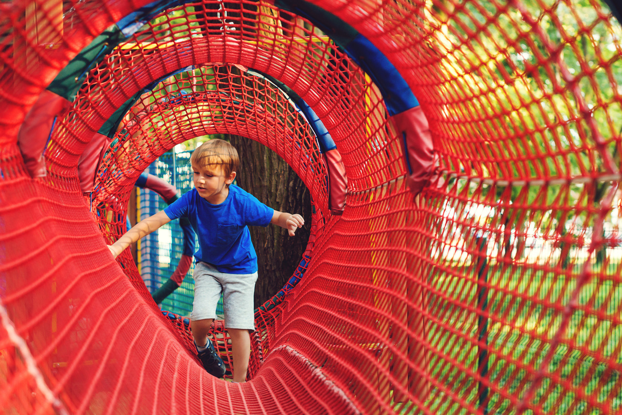 Happy kid overcomes obstacles in rope adventure park. Summer holidays concept. Little boy playing at rope adventure park. Modern amusement park for kids. Outdoors games